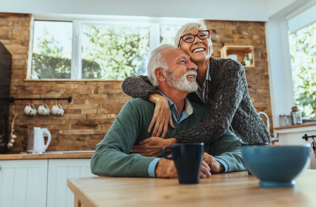 An older adult hugs their spouse while settling into their new kitchen during breakfast time in senior living