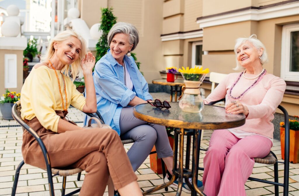 3 older friends laugh together while sitting at an antique glass patio table outdoors in a courtyard