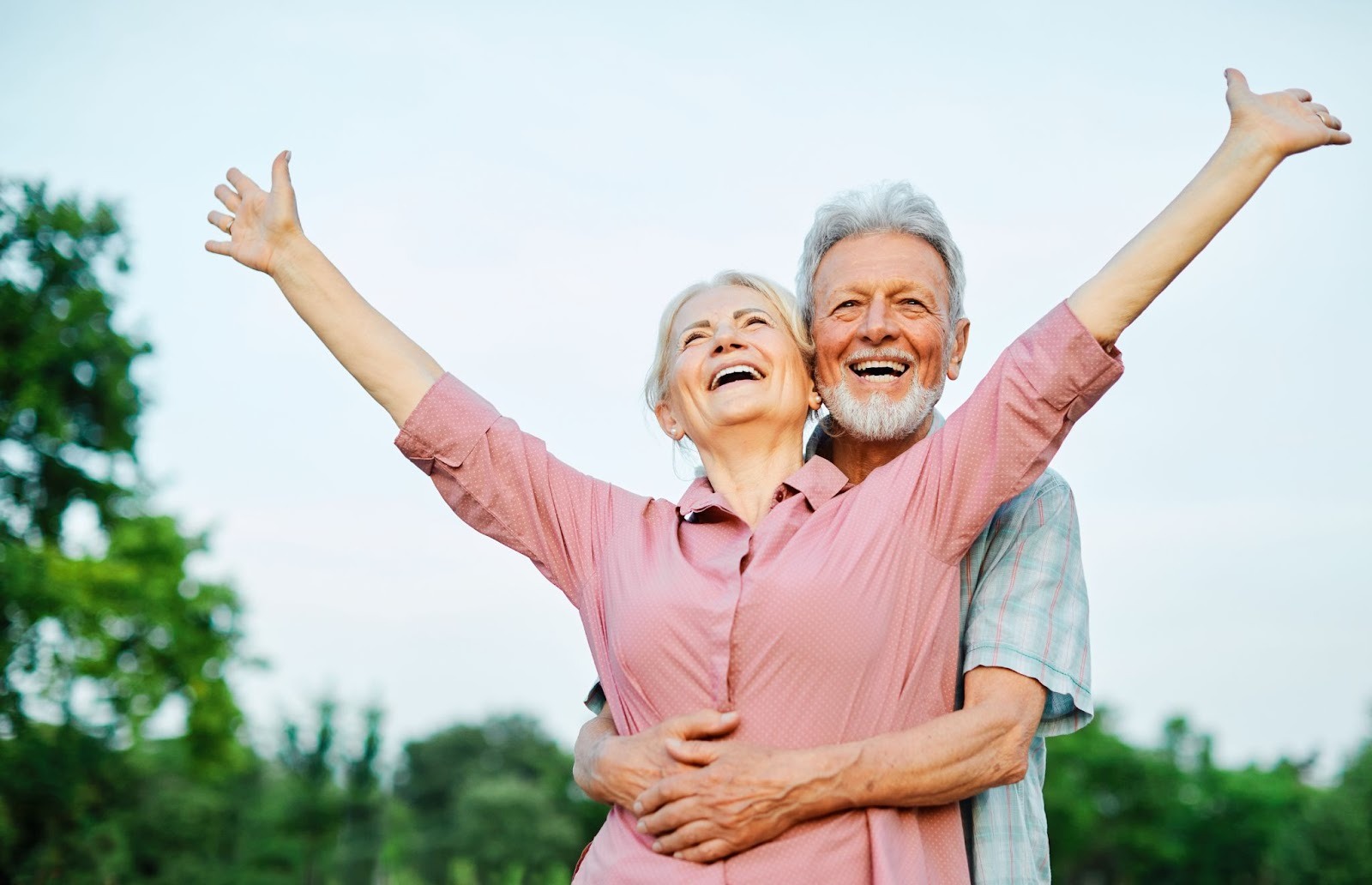 An older adult celebrates and raises their arm while their smiling spouse hugs them from behind